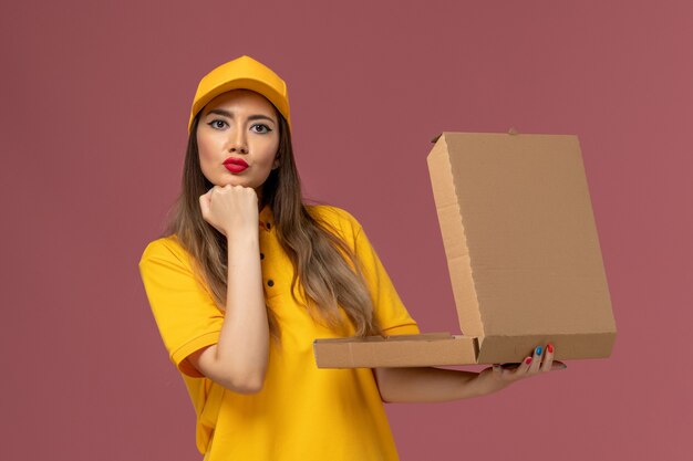 Vue avant du courrier féminin en uniforme jaune et cap tenant la boîte de nourriture ouverte sur le mur rose clair