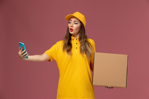 Vue avant du courrier féminin en uniforme jaune et cap tenant la boîte de nourriture et téléphone de travail sur mur rose clair