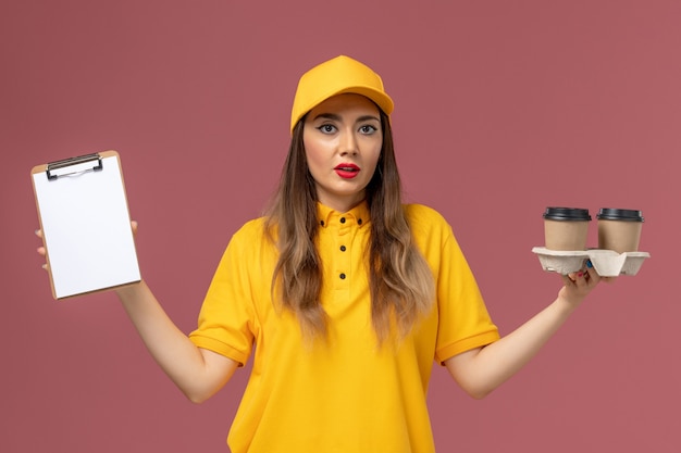 Photo gratuite vue avant du courrier féminin en uniforme jaune et cap tenant la livraison tasses à café et bloc-notes sur mur rose clair