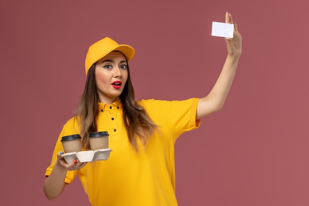 Vue avant du courrier féminin en uniforme jaune et cap tenant la livraison tasses à café et carte sur mur rose