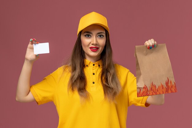 Vue avant du courrier féminin en uniforme jaune et cap tenant le paquet alimentaire et carte en plastique sur le mur rose