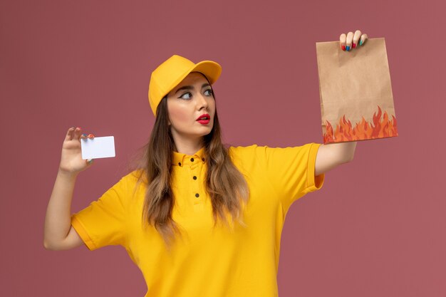 Vue avant du courrier féminin en uniforme jaune et cap tenant le paquet alimentaire et carte en plastique sur le mur rose