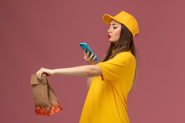 Vue avant du courrier féminin en uniforme jaune et cap tenant le paquet alimentaire et prendre une photo sur le mur rose