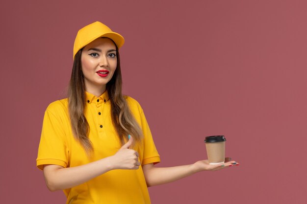 Vue avant du courrier féminin en uniforme jaune et cap tenant la tasse de café de livraison sur le mur rose