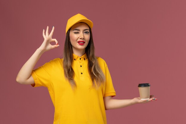 Vue avant du courrier féminin en uniforme jaune et cap tenant la tasse de café de livraison sur le mur rose