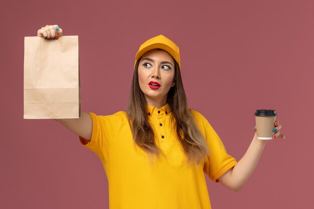 Vue avant du courrier féminin en uniforme jaune et cap tenant la tasse de café de livraison et le paquet de nourriture sur le mur rose