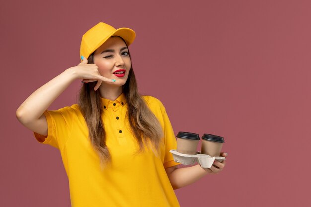 Vue avant du courrier féminin en uniforme jaune et cap tenant des tasses de café de livraison sur le mur rose