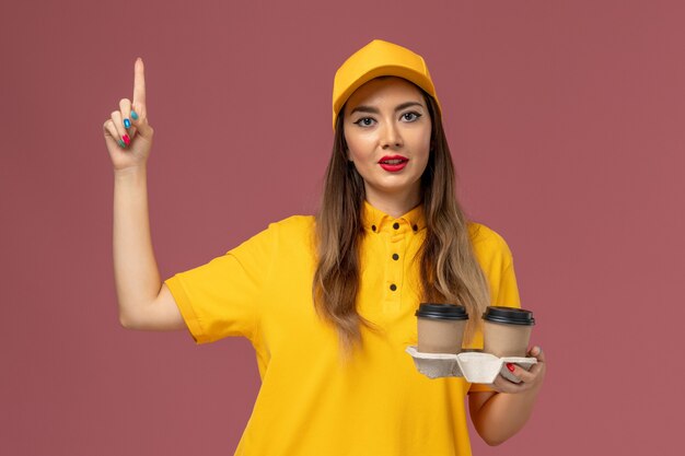 Vue avant du courrier féminin en uniforme jaune et cap tenant des tasses de café de livraison sur le mur rose