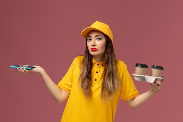 Vue avant du courrier féminin en uniforme jaune et cap tenant des tasses de café de livraison et smartphone sur bureau rose