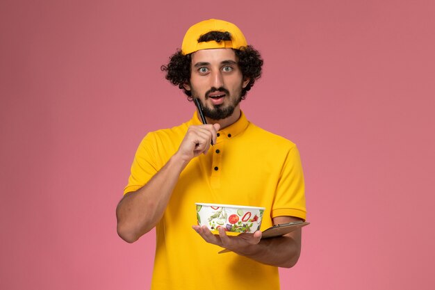 Vue avant du courrier masculin en cape uniforme jaune avec bol de livraison rond sur ses mains en écrivant des notes sur le fond rose.