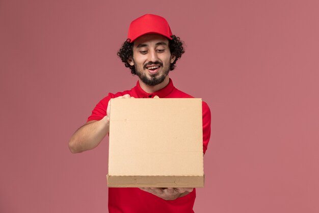 Vue avant du courrier masculin en chemise rouge et cape tenant la boîte de nourriture de livraison sur le mur rose clair