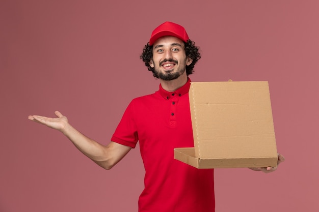 Vue avant du courrier masculin en chemise rouge et cape tenant la boîte de nourriture de livraison sur le mur rose