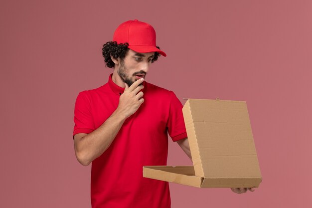 Vue avant du courrier masculin en chemise rouge et cape tenant la boîte de nourriture de livraison vide sur le mur rose clair