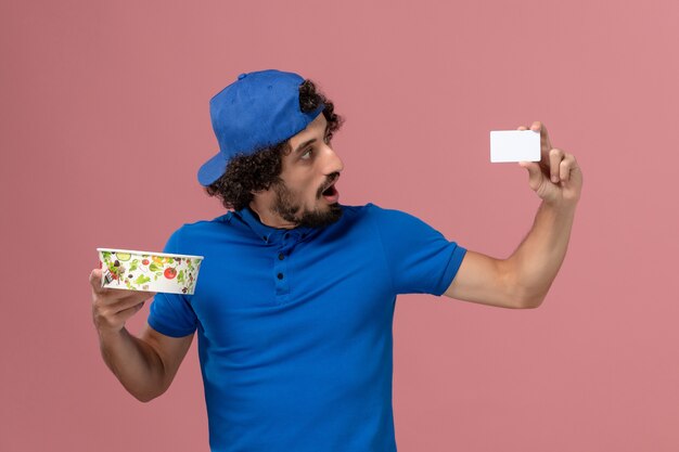 Vue avant du courrier masculin en uniforme bleu et cape tenant le bol et la carte de livraison ronde sur mur rose