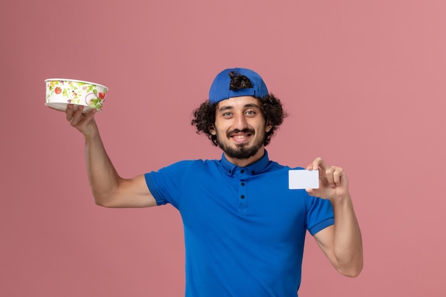 Vue avant du courrier masculin en uniforme bleu et cape tenant le bol et la carte de livraison ronde sur mur rose