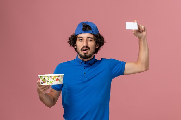 Vue avant du courrier masculin en uniforme bleu et cape tenant le bol et la carte de livraison ronde sur mur rose