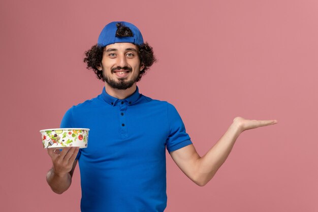 Vue avant du courrier masculin en uniforme bleu et cape tenant un bol de livraison rond sur le mur rose