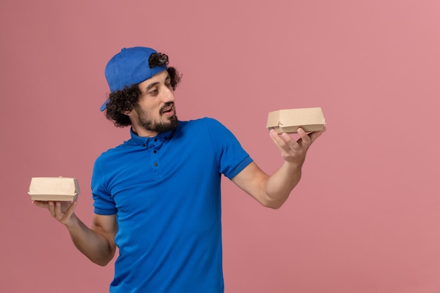 Vue avant du courrier masculin en uniforme bleu et cape tenant peu de colis alimentaires de livraison sur le mur rose