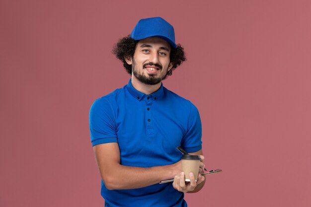 Vue avant du courrier masculin en uniforme bleu et capuchon avec tasse de café de livraison et bloc-notes sur ses mains en écrivant des notes sur le mur rose