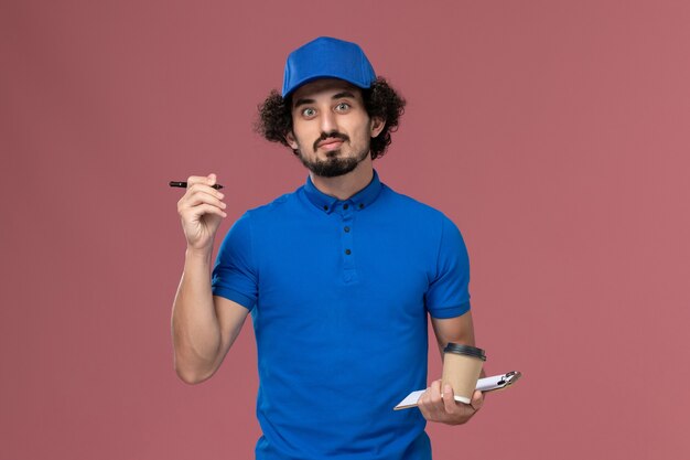 Vue avant du courrier masculin en uniforme bleu et capuchon avec tasse de café de livraison et bloc-notes sur ses mains en écrivant des notes sur le mur rose