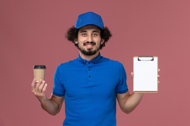 Vue avant du courrier masculin en uniforme bleu et capuchon avec tasse de café de livraison et bloc-notes sur ses mains sur le mur rose