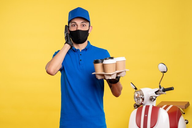 Vue avant du courrier masculin en uniforme bleu et masque tenant du café sur le service d'emploi uniforme jaune - pandémie de livraison de covid