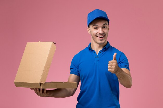 Vue avant du courrier masculin en uniforme bleu tenant la boîte de nourriture souriant sur le mur rose, la prestation de services uniforme de travailleur d'emploi