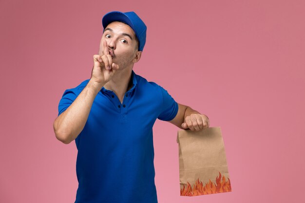 Vue avant du courrier masculin en uniforme bleu tenant le paquet alimentaire demandant de se taire sur le mur rose, la prestation de services uniforme de travailleur d'emploi