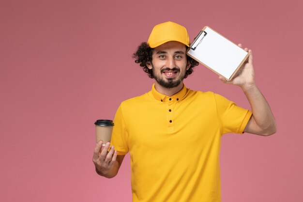 Vue avant du courrier masculin en uniforme jaune et cape tenant la tasse de café de livraison et le bloc-notes sur le mur rose