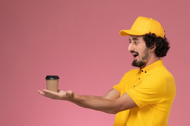 Vue avant du courrier masculin en uniforme jaune et cape tenant la tasse de café de livraison sur le mur rose clair