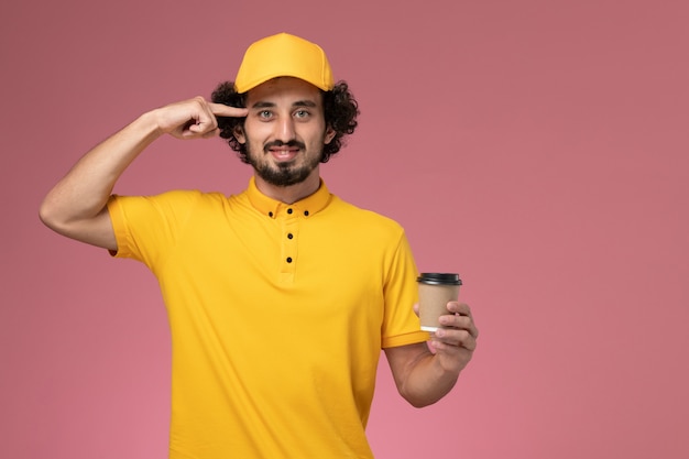 Vue avant du courrier masculin en uniforme jaune et cape tenant la tasse de café de livraison sur le mur rose clair