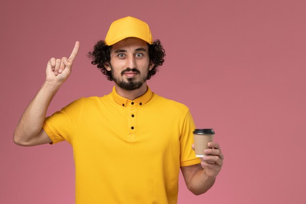 Vue avant du courrier masculin en uniforme jaune et cape tenant la tasse de café de livraison sur le mur rose clair