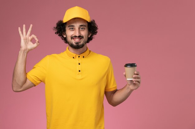 Vue avant du courrier masculin en uniforme jaune et cape tenant la tasse de café de livraison sur le mur rose