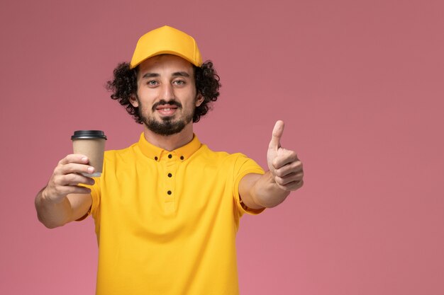 Vue avant du courrier masculin en uniforme jaune et cape tenant la tasse de café de livraison sur le mur rose