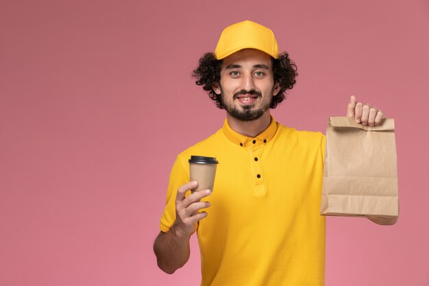 Vue avant du courrier masculin en uniforme jaune et cape tenant la tasse de café de livraison et le paquet de nourriture sur le mur rose