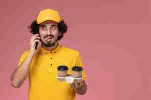 Photo gratuite vue avant du courrier masculin en uniforme jaune et cape tenant des tasses de café de livraison marron parler au téléphone sur mur rose