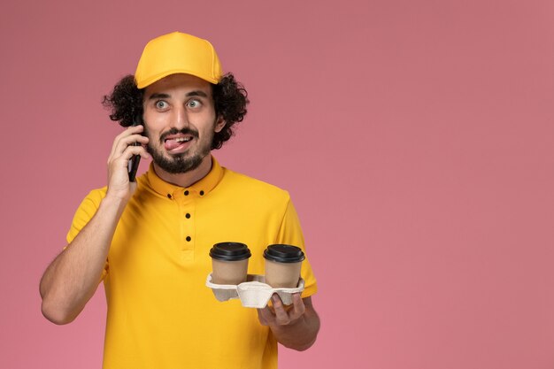 Vue avant du courrier masculin en uniforme jaune et cape tenant des tasses de café de livraison marron parler au téléphone sur mur rose