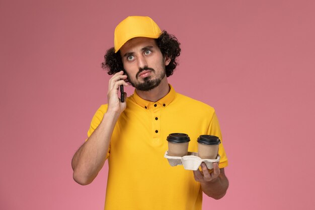 Vue avant du courrier masculin en uniforme jaune et cape tenant des tasses de café de livraison marron parler au téléphone sur mur rose