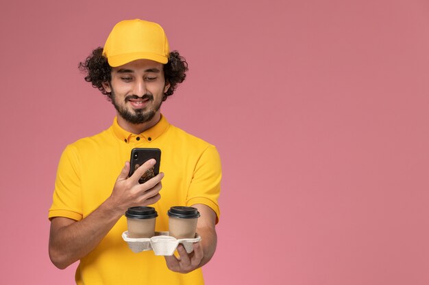 Vue avant du courrier masculin en uniforme jaune et cape tenant des tasses de café de livraison marron et en prenant une photo d'eux sur le mur rose