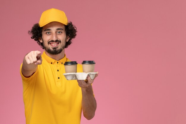 Vue avant du courrier masculin en uniforme jaune et cape tenant des tasses de café de livraison marron avec un sourire sur le mur rose