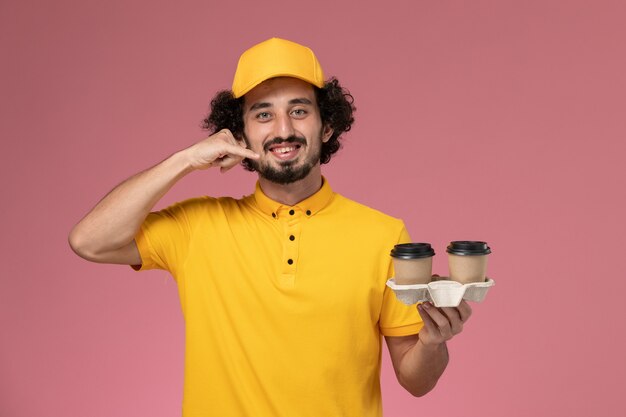 Vue avant du courrier masculin en uniforme jaune et cape tenant des tasses de café de livraison sur le mur rose