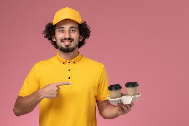 Vue avant du courrier masculin en uniforme jaune et cape tenant des tasses de café de livraison sur le mur rose