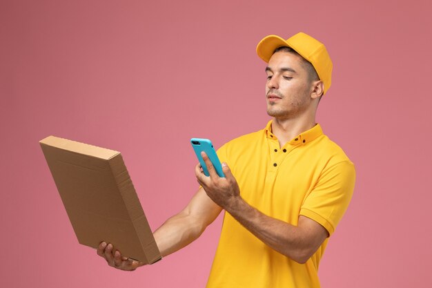 Vue avant du courrier masculin en uniforme jaune en prenant une photo de la boîte de livraison de nourriture sur bureau rose