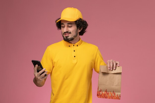 Vue avant du courrier masculin en uniforme jaune tenant le paquet alimentaire parler au téléphone sur le mur rose