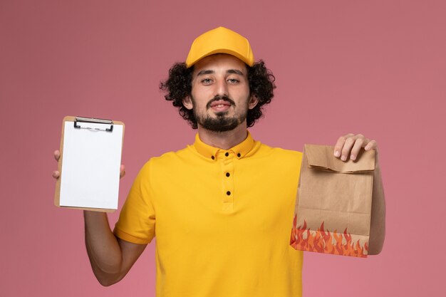 Vue avant du courrier masculin en uniforme jaune tenant le paquet de nourriture et le bloc-notes sur le mur rose