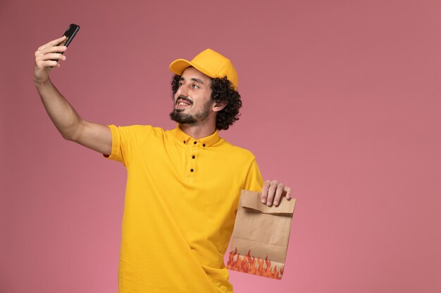 Vue avant du courrier masculin en uniforme jaune tenant le paquet de nourriture et en prenant une photo sur le mur rose