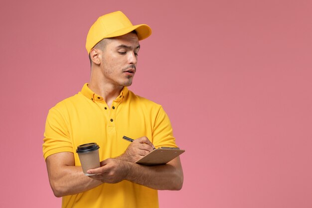 Vue avant du courrier masculin en uniforme jaune tenant la tasse de café de livraison et le bloc-notes écrivant des notes sur le bureau rose
