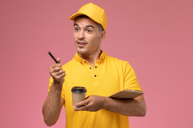 Vue avant du courrier masculin en uniforme jaune tenant la tasse de café de livraison et le bloc-notes écrivant des notes sur le bureau rose