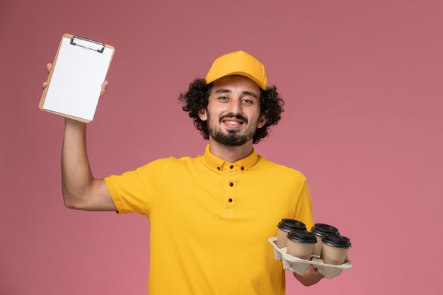 Vue avant du courrier masculin en uniforme jaune tenant des tasses de café de livraison marron et bloc-notes sur mur rose