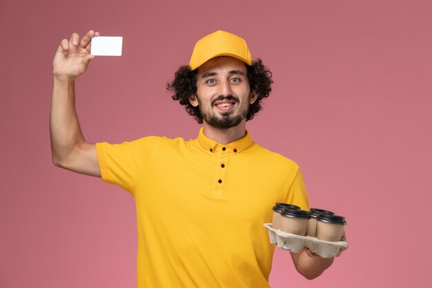 Vue avant du courrier masculin en uniforme jaune tenant des tasses de café de livraison marron et carte blanche sur mur rose clair